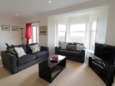 A living room with two grey sofas and a black coffee table facing a television by bay windows at Apartment 3 Marian Y Mor in Pwllheli