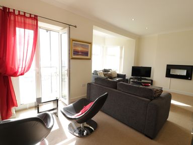 A living room with grey sofas black chairs red curtains a wall-mounted tv and a painting at Apartment 3 Marian Y Mor in Pwllheli