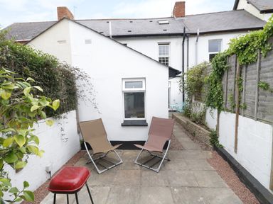 A small outdoor patio area with two lounge chairs and a red barbecue grill surrounded by white walls and greenery at No 9 in Watchet