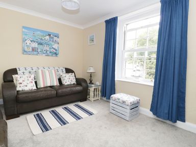 A living room with a brown sofa with cushions and a small table with a lamp beside a window with blue curtains at No 9 in Watchet