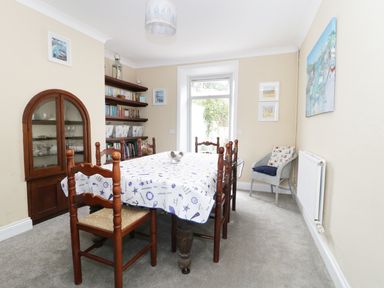 A dining room with a wooden table covered by a patterned tablecloth surrounded by wooden chairs a glass-fronted cabinet shelves with books and decorative items a gray armchair with cushions and framed pictures on the walls at No 9 in Watchet