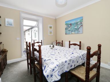 A dining room with a wooden table and six chairs a white and blue tablecloth and paintings on the walls at No 9 in Watchet