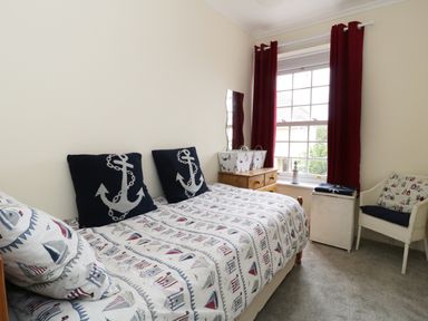 A bedroom with a single bed covered with nautical themed bedding and pillows next to a window with red curtains and a wooden dresser at No 9 Watchet