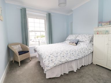 A bedroom with a bed with patterned bedding a wicker chair by a window with curtains and a white cabinet at No 9 Watchet