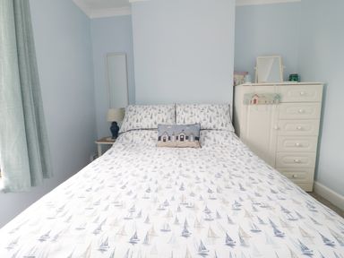 A bedroom with a bed featuring sailboat patterned bedding a side table with a lamp a tall mirror and a white dresser with a small mirror at No 9 Watchet