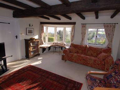 A living room with exposed wooden beams a red patterned sofa and a large window with a curved bench at Appletree Cottage in Higher Brimley near Bovey Tracey