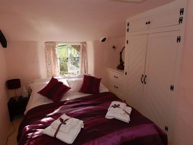 A bedroom with a bed covered in purple bedding two sets of white towels on the bed a window with floral curtains a white built-in cupboard and dresser with mirror at Appletree Cottage in Higher Brimley near Bovey Tracey