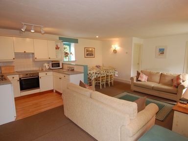 An open plan living room with two beige sofas a green rug a wooden dining table with chairs and a kitchen area with white cabinets at The Coach House in Ugborough
