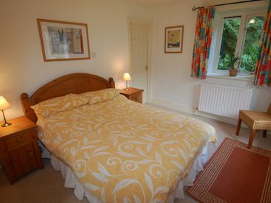 A bedroom with a double bed two bedside tables with lamps framed pictures a window with curtains and a chair at The Coach House in Ugborough