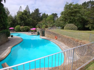An outdoor swimming pool with stone walls and pink umbrellas near trees at The Coach House in Ugborough