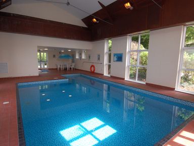 An indoor swimming pool with blue tiles white plastic chairs and table under a blue umbrella at The Coach House in Ugborough