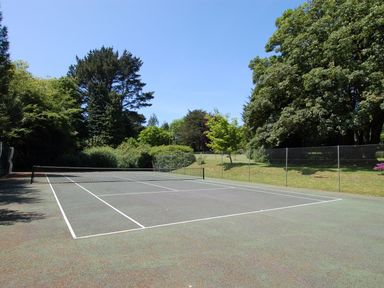 An outdoor tennis court surrounded by green trees and a chain link fence at The Coach House in Ugborough
