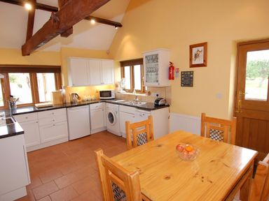 A kitchen with white cabinets wooden dining table and chairs and a door at The Milking Parlour in Wookey