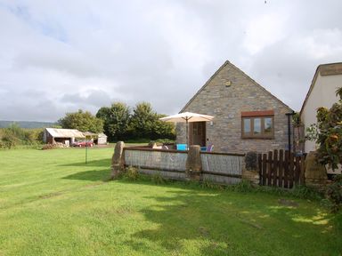 A stone house with an outdoor table and umbrella on a grass lawn with a small shed and trees in the background at The Milking Parlour in Wookey