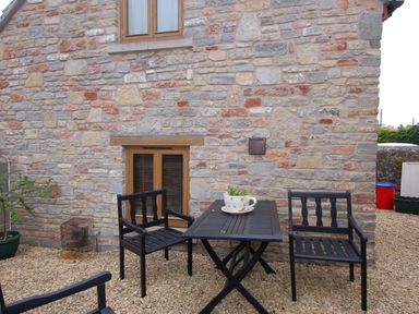 An outdoor patio area with a black table and three black chairs on gravel next to a stone wall with two windows at The Old Cider Barn in Wookey