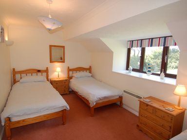 A bedroom with two single beds a window with striped valance and wooden furniture at Pine Cottage in Maenporth