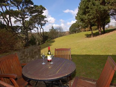 A wooden outdoor table with wine bottle and glasses on green lawn with trees and bushes at Pine Cottage in Maenporth