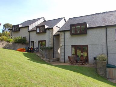 A two story house with two outdoor seating areas on a grassy slope at Pine Cottage in Maenporth