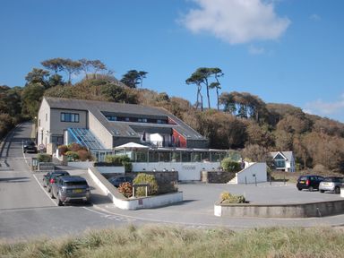 A building with a parking lot and several parked cars near a wooded hill under a blue sky at Pine Cottage in Maenporth
