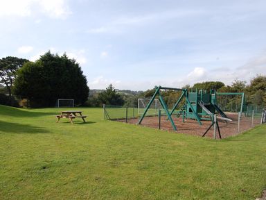 An outdoor playground with swings slides and fenced area next to a picnic table and soccer goals on a grassy field at Pine Cottage in Maenporth