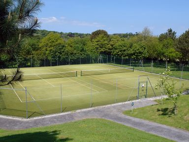 An outdoor tennis court with a green playing surface surrounded by a chain-link fence and trees at Pine Cottage in Maenporth