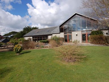 A building with large windows surrounded by bushes and a lawn with trees and picnic tables at Pine Cottage in Maenporth