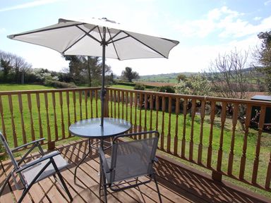 A wooden deck with a glass table and two chairs under a large patio umbrella overlooking a grassy yard at Little Waycroft in Dawlish