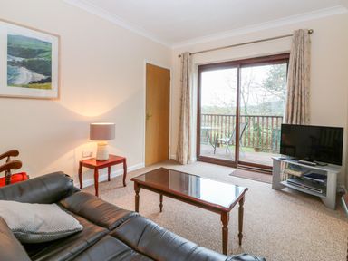 A living room with a leather sofa a wooden coffee table a lamp on a side table a television on a stand and sliding glass doors leading to a balcony at Little Waycroft in Dawlish