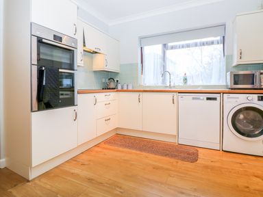 A kitchen with white cabinets, built-in oven, dishwasher, washing machine, microwave, and a window with a sink below at Little Waycroft in Dawlish