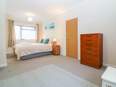 A bedroom with a bed side table with a lamp a wooden dresser and a window with curtains at Little Waycroft in Dawlish