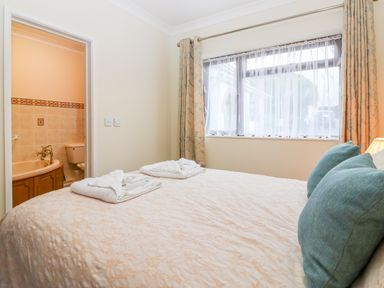 A bedroom with a double bed and blue pillows next to a window with curtains and an ensuite bathroom with a bathtub at Little Waycroft in Dawlish