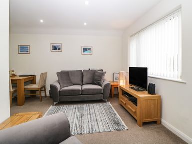 A living room with a gray sofa, wooden TV stand with a TV, a lamp, a rug, a wooden dining table with chairs, and pictures on the wall at Monkbarns in Brixham