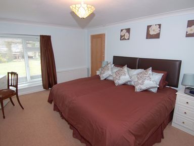 A bedroom with a large bed with brown bedding and multiple pillows bedside tables with lamps a chair and a window with brown curtains at Garden View in Kingskerswell
