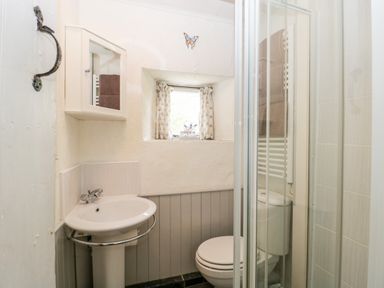 A small bathroom with a white sink pedestal, toilet, shower cubicle, small window with curtains and a wall cabinet at The Granary in Edington near Street and Glastonbury