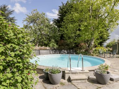 An outdoor round swimming pool with metal handrails surrounded by plants and trees with a small table and chairs nearby at The Granary in Edington near Street and Glastonbury