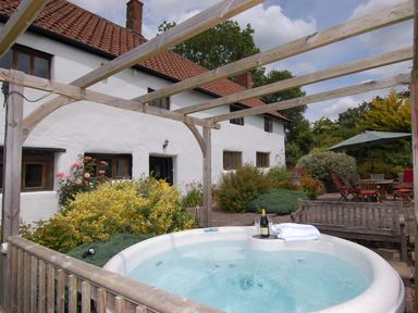 An outdoor area with a hot tub surrounded by wooden beams a garden with shrubs and flowers wooden benches and a patio with a table and chairs at Surridge Farmhouse in Waterrow