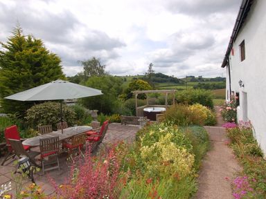 An outdoor patio area with table and chairs under an umbrella and a garden pathway next to a white building at Surridge Farmhouse in Waterrow