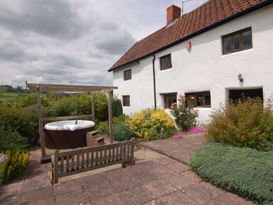 An outdoor patio with a hot tub under a wooden frame and a wooden bench near a white house with plants at Surridge Farmhouse in Waterrow