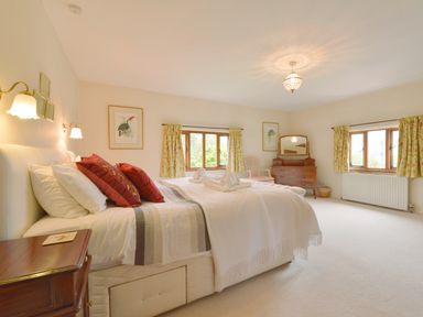 A bedroom with a bed with red pillows two windows with curtains a wooden dresser with a mirror and a chair at Surridge Farmhouse in Waterrow