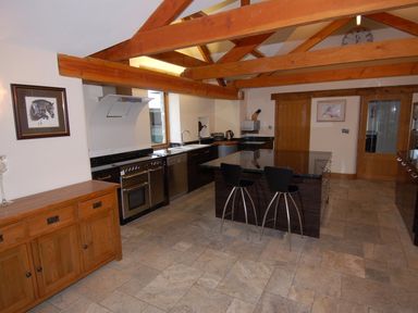 A kitchen with wooden beams tiled floor black countertop island with two stools wooden cabinets and framed pictures at Hideaway in Tintagel
