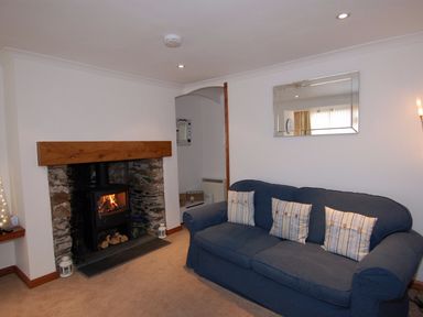 A living room with a blue sofa and three striped cushions a fireplace with burning wood and stone surround and a large mirror on the wall at The Keep Pot in Port Isaac