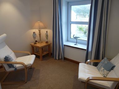 A small room with two white cushioned wooden chairs a wooden side table with a lamp and decorations and a window with curtains at The Keep Pot in Port Isaac