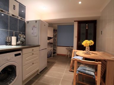 A kitchen with washing machine fridge wooden table and chairs with yellow flowers at The Keep Pot in Port Isaac