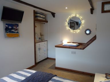 A bedroom with a wall-mounted television striped bedding a dresser an open closet and a decorative mirror with lights at The Keep Pot in Port Isaac