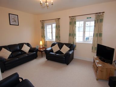 A living room with black leather sofas a wooden TV stand with a television and windows with curtains at The Barn in Tregoodwell