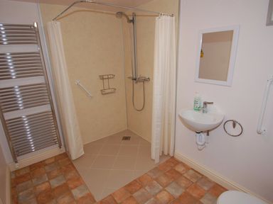 A bathroom with a corner shower area with white curtains a small wall-mounted sink with a soap dispenser a mirror a towel rack and tile flooring at The Barn in Tregoodwell
