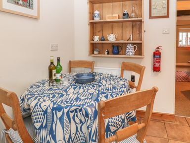 A small dining area with a round table with blue and white fish patterned tablecloth four wooden chairs a bottle of wine and a bottle of liqueur two glasses a blue bowl and wooden shelves with various kitchenware and a fire extinguisher on the wall at Robyn Cottage in Otterton