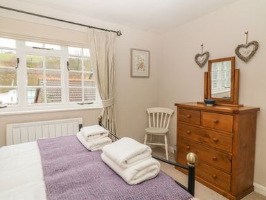 A bedroom with a bed, folded towels, a wooden dresser with mirror, a chair, and a window with curtains at Robyn Cottage in Otterton