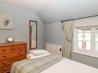 A bedroom with a wooden chest of drawers a mirror a bed with folded towels and a window with curtains at Robyn Cottage in Otterton