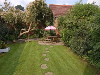 A garden with a wooden picnic table and pink umbrella surrounded by trees and bushes at Robyn Cottage in Otterton